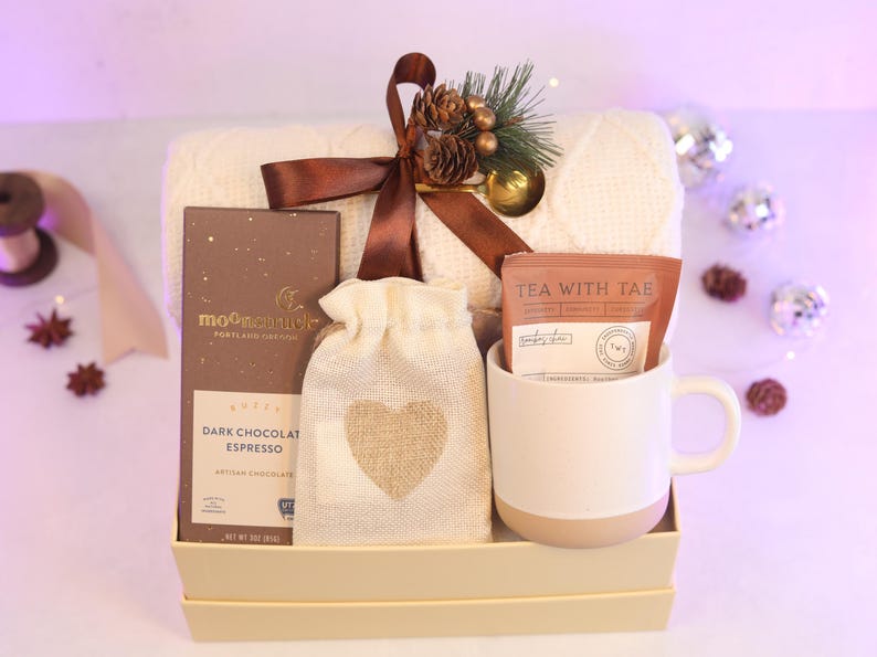 May include: A gift set in a box containing a cream-colored cable knit blanket tied with a brown ribbon, a bar of Moonstruck dark chocolate espresso, a small beige drawstring pouch with a burlap heart, and a white ceramic mug with a tea packet.