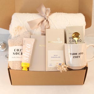 May include: A brown cardboard gift box filled with cozy items for a self-care night. The box contains a white knitted blanket, a pair of fuzzy socks, a tube of shea butter lotion, a notebook, a bar of milk chocolate, a mug with the text "Warm and Cozy", and a gold spoon.