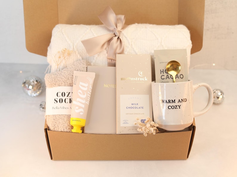 May include: A brown cardboard gift box filled with cozy items for a self-care night. The box contains a white knitted blanket, a pair of fuzzy socks, a tube of shea butter lotion, a notebook, a bar of milk chocolate, a mug with the text "Warm and Cozy", and a gold spoon.