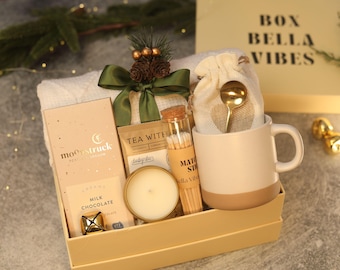 May include: A gift box labeled BOX BELLA VIBES containing a Moonstruck milk chocolate bar, a tea packet, a candle in a gold tin, a glass vial of matches, a cream-colored mug with a gold spoon in a fabric pouch, and a green ribbon.