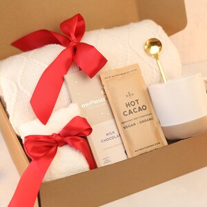 May include: A gift box containing a white textured throw blanket with a red ribbon, a Moonstruck milk chocolate bar, a packet of Retreat Drinks hot cacao, a gold-toned spoon, and a white and beige ceramic mug with a speckled finish.