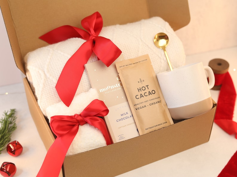 May include: A gift box containing a white textured throw blanket with a red ribbon, a Moonstruck milk chocolate bar, a packet of Retreat Drinks hot cacao, a gold-toned spoon, and a white and beige ceramic mug with a speckled finish.