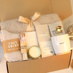 May include: A gift box filled with cosy items for relaxation, including a blanket, socks, a candle, hot chocolate mix, a mug, and matches. The box is decorated with a gold ribbon and the words "Cozy Socks" and "Mom" are visible.