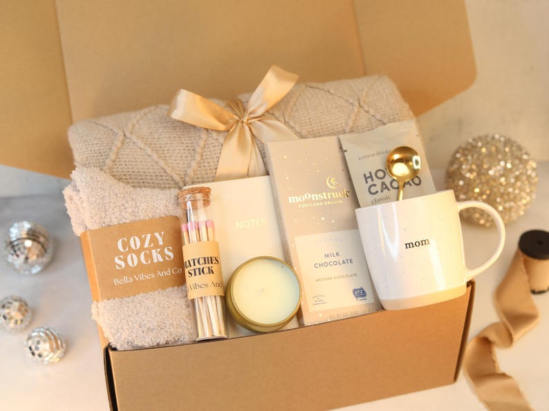 May include: A gift box filled with cosy items for relaxation, including a blanket, socks, a candle, hot chocolate mix, a mug, and matches. The box is decorated with a gold ribbon and the words "Cozy Socks" and "Mom" are visible.