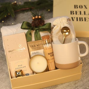 May include: A gift box containing a Moonstruck milk chocolate bar, a small candle, a glass tube of matchsticks, a beige mug with a gold spoon in a fabric pouch, and a white knit item, all decorated with a green ribbon and pine cones.