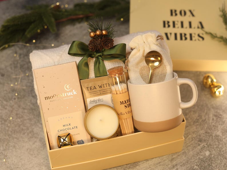 May include: A gift box containing a Moonstruck milk chocolate bar, a small candle, a glass tube of matchsticks, a beige mug with a gold spoon in a fabric pouch, and a white knit item, all decorated with a green ribbon and pine cones.