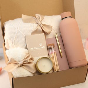 May include: A gift box filled with self-care items, including a white blanket, a candle, a water bottle, matches, a notebook, and a pen. The box is decorated with a beige ribbon and bow.