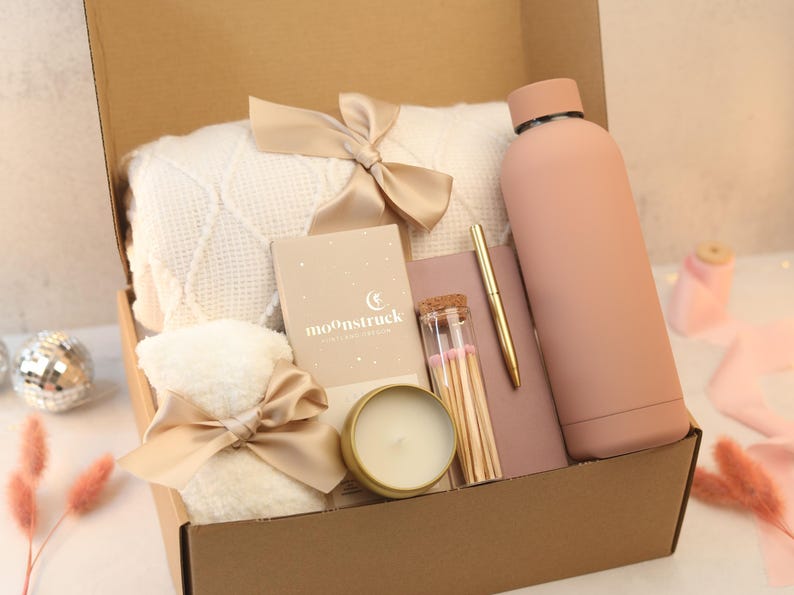 May include: A gift box filled with self-care items, including a white blanket, a candle, a water bottle, matches, a notebook, and a pen. The box is decorated with a beige ribbon and bow.