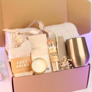 Puede incluir: Una caja de regalo que contiene una manta de punto beige con cinta, un par de calcetines suaves con la etiqueta Cozy Socks, una barra de chocolate Moonstruck, una vela peque&ntilde;a, un frasco de cerillas, un bol&iacute;grafo dorado, un cuaderno y un vaso dorado.