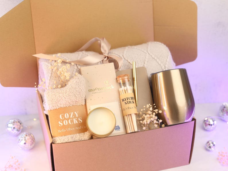 Puede incluir: Una caja de regalo que contiene una manta de punto beige con cinta, un par de calcetines suaves con la etiqueta Cozy Socks, una barra de chocolate Moonstruck, una vela peque&ntilde;a, un frasco de cerillas, un bol&iacute;grafo dorado, un cuaderno y un vaso dorado.