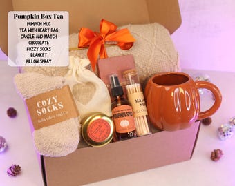 Op de afbeelding: Een geschenkdoos met het label Pumpkin Box Tea bevat een pompoenvormige mok, een beige gebreide deken met een oranje lint, pluizige sokken, een klein zakje met een hartvormige uitsparing, een chocoladereep, een glazen buisje met lucifers en spray.