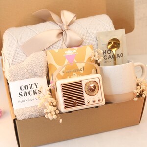 Puede incluir: Una caja de regalo llena de artículos acogedores. Incluye una manta color crema atada con un lazo, un par de calcetines suaves, una pequeña radio, una taza con una cuchara dorada y una barra de caramelo. La caja está decorada con adornos rosas.