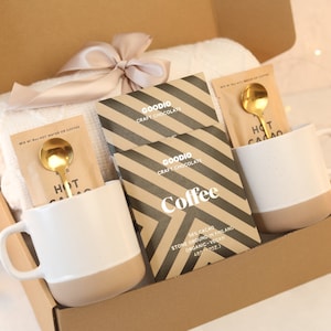 May include: A brown cardboard box filled with a cosy gift set. The set includes two white and beige mugs, two gold spoons, two packets of hot chocolate mix, two packets of craft chocolate, and a white throw blanket. The box is tied with a beige ribbon.