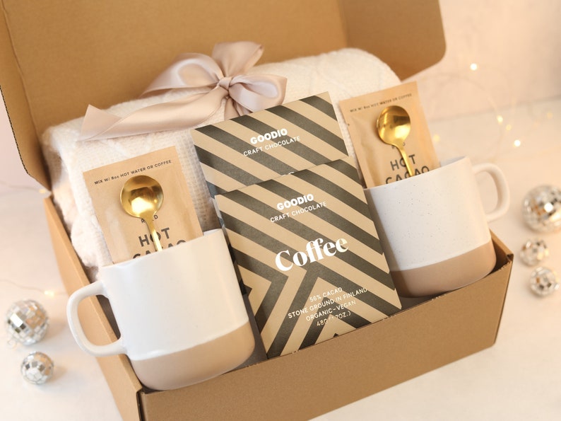 May include: A brown cardboard box filled with a cosy gift set. The set includes two white and beige mugs, two gold spoons, two packets of hot chocolate mix, two packets of craft chocolate, and a white throw blanket. The box is tied with a beige ribbon.