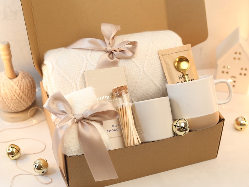 May include: A gift box filled with cozy items for a relaxing evening. It includes a white blanket, a white towel, a mug, a small jar of matches, a packet of hot chocolate, and a gold spoon. The box is decorated with a brown ribbon and gold bells.