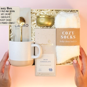 May include: A gift box containing a two-toned speckled mug with a gold spoon, a packet of hot cacao, a gold-toned candle tin, a Moonstruck milk chocolate bar, and a pair of white fuzzy socks with a label reading Cozy Socks Bella Vibes And Co.