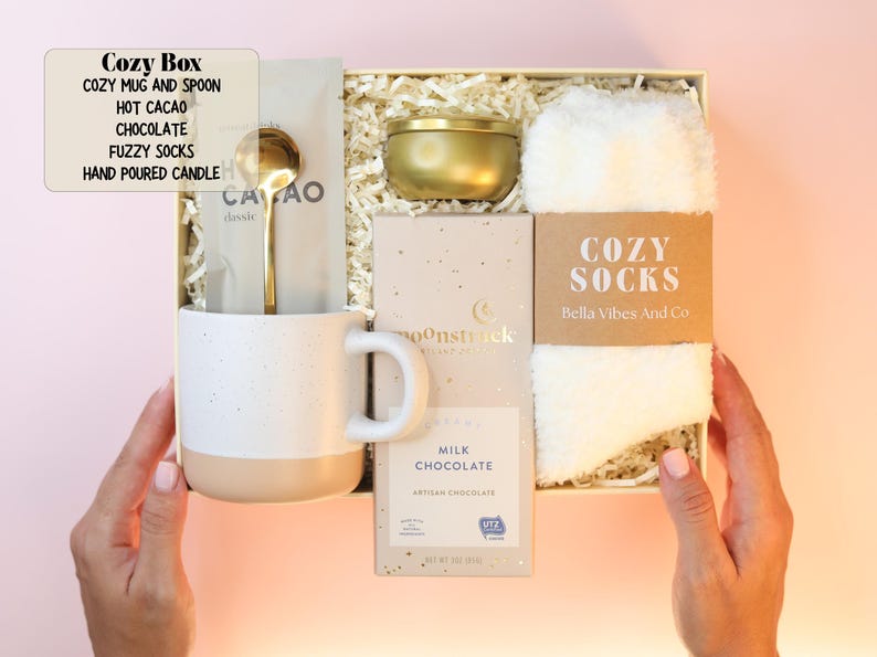 May include: A gift box containing a two-toned speckled mug with a gold spoon, a packet of hot cacao, a gold-toned candle tin, a Moonstruck milk chocolate bar, and a pair of white fuzzy socks with a label reading Cozy Socks Bella Vibes And Co.
