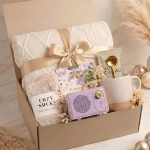 May include: A gift box containing a cream-colored cable knit blanket tied with a gold ribbon, a pair of beige fuzzy socks labeled COZY SOCKS, a lavender-colored vintage-style radio, a ceramic mug, a gold spoon, and a packet labeled HOT CACAO.