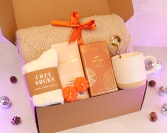 May include: A gift box containing a beige cable-knit blanket tied with an orange ribbon, a pair of white fuzzy socks labeled Cozy Socks, a milk chocolate bar, a box of salted caramel hot cocoa, two orange pumpkin-shaped candles, and a mug with a spoon.