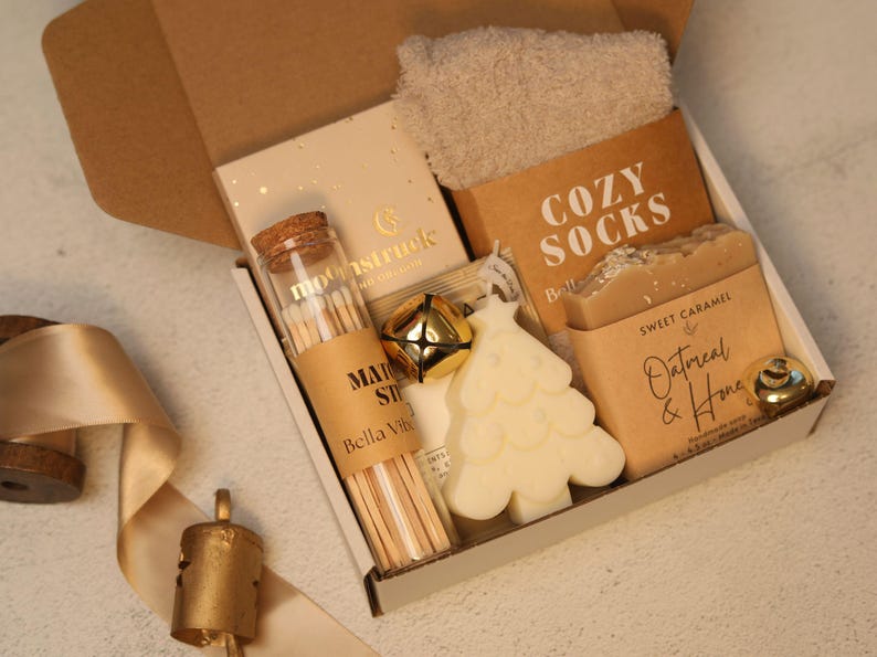 May include: A gift box containing a pair of beige fuzzy socks, a glass vial of matches with a cork stopper, a gold jingle bell, a white Christmas tree-shaped candle, and a bar of Sweet Caramel Oatmeal & Honey soap with brown paper labels.