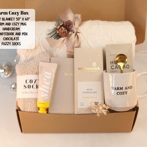 May include: A gift box containing a 50" x 60" white knit blanket, a speckled ceramic mug with the text "WARM AND COZY", a tube of shea butter hand cream, a beige A6 notebook with a pen, a Moonstruck milk chocolate bar, and fuzzy socks.
