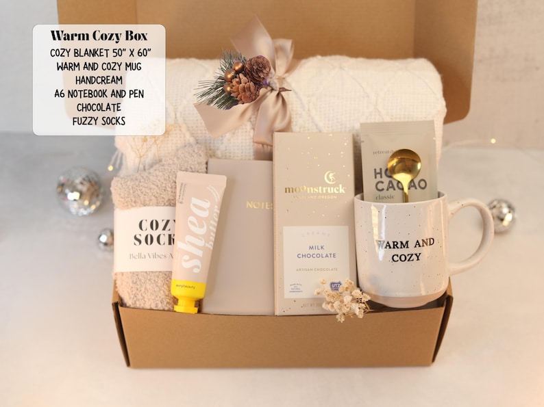 May include: A gift box containing a 50" x 60" white knit blanket, a speckled ceramic mug with the text "WARM AND COZY", a tube of shea butter hand cream, a beige A6 notebook with a pen, a Moonstruck milk chocolate bar, and fuzzy socks.