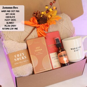Puede incluir: Una caja de regalo con temática otoñal llena de artículos acogedores. Incluye una taza, chocolate caliente, chocolate, calcetines afelpados, una manta, spray para almohadas y pino de hojas de otoño. La taza dice "Warm and Cozy".