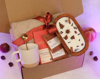 May include: A gift box filled with a beige knit blanket, a white and beige mug with a gold spoon, chocolate bars, and a wooden candle holder with a white candle decorated with spices and flowers. A red bow adorns the box.