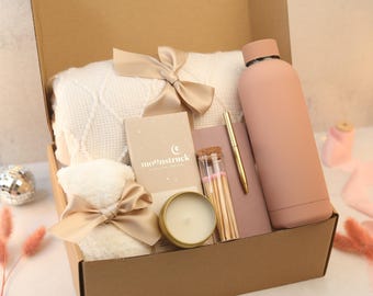May include: A gift box containing a cream-coloured textured blanket tied with a champagne ribbon, a matte dusty rose water bottle, a Moonstruck chocolate bar, a small gold-rimmed candle, a glass vial of matches, a dusty rose notebook, and a gold pen.