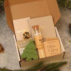May include: A gift box containing a beige notebook labeled NOTES, a glass vial of matches labeled Bella Villa, a bar of Sweet Caramel Oatmeal & Honey soap, a green tree-shaped candle, a gold bell, and a tea packet labeled TEA WITH.