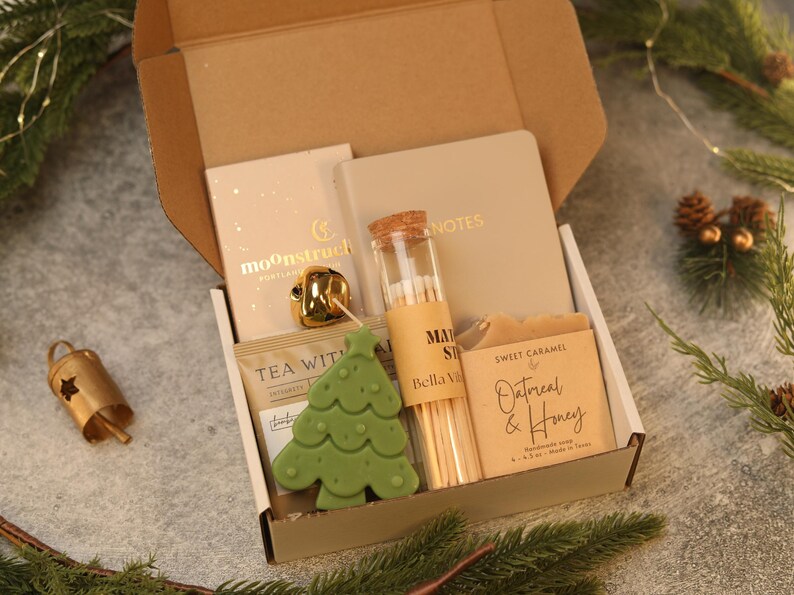 May include: A gift box containing a beige notebook labeled NOTES, a glass vial of matches labeled Bella Villa, a bar of Sweet Caramel Oatmeal & Honey soap, a green tree-shaped candle, a gold bell, and a tea packet labeled TEA WITH.