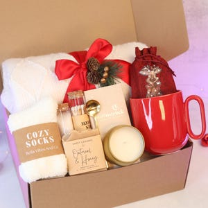 Puede incluir: Una caja de regalo que contiene una manta de punto blanca con un lazo rojo, calcetines blancos difusos con la etiqueta Cozy Socks, un vial de cerillas, una cuchara dorada, jab&oacute;n de avena y miel, una vela y una taza roja.