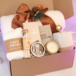 Puede incluir: Una caja de regalo que contiene una manta de punto blanca con una cinta marr&oacute;n, calcetines blancos suaves con la etiqueta Cozy Socks, un vial de vidrio con cerillas, una barra de chocolate Moonstruck, un sobre de cacao, una cuchara dorada, una taza blanca, una vela y un adorno de madera con forma de ciervo.