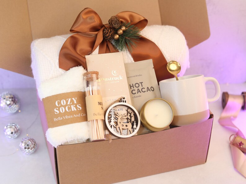 Puede incluir: Una caja de regalo que contiene una manta de punto blanca con una cinta marr&oacute;n, calcetines blancos suaves con la etiqueta Cozy Socks, un vial de vidrio con cerillas, una barra de chocolate Moonstruck, un sobre de cacao, una cuchara dorada, una taza blanca, una vela y un adorno de madera con forma de ciervo.