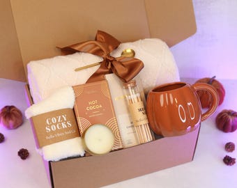 May include: A gift box containing a white textured blanket tied with a brown ribbon, a pair of white fuzzy socks labeled Cozy Socks, a box of hot cocoa, a small candle, a glass vial of matches, and an orange pumpkin-shaped ceramic mug.