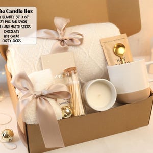 May include: A gift box containing a 50 inch by 60 inch white knit blanket, a two-tone mug with a gold spoon, a candle, a glass jar of matchsticks, a chocolate bar, hot cacao mix, and fuzzy socks, all tied with a beige satin ribbon.