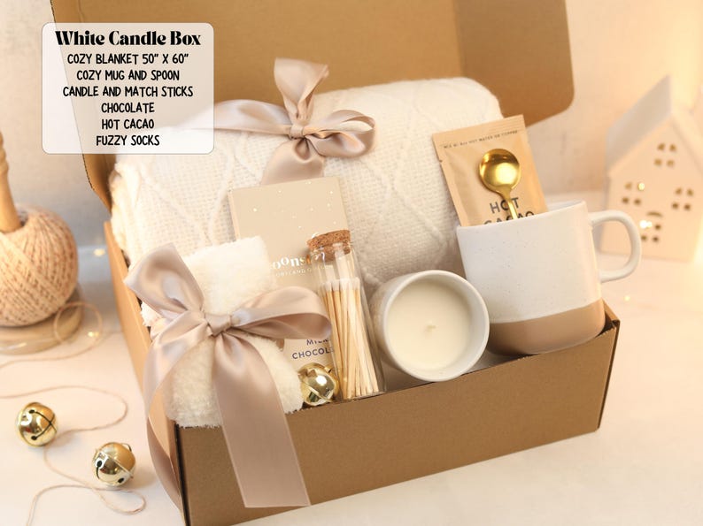 May include: A gift box containing a 50 inch by 60 inch white knit blanket, a two-tone mug with a gold spoon, a candle, a glass jar of matchsticks, a chocolate bar, hot cacao mix, and fuzzy socks, all tied with a beige satin ribbon.