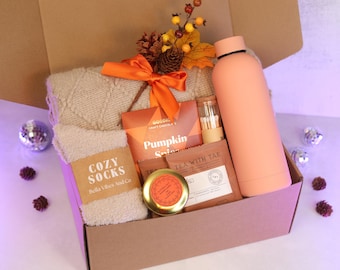 May include: A gift box filled with cozy items. Includes a peach-colored water bottle, a pair of beige socks labeled "Cozy Socks", and a selection of tea and chocolate. The box is decorated with an orange bow and fall foliage.