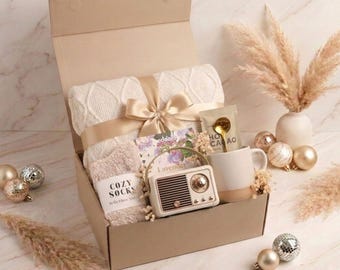 May include: A gift box containing a cream-colored cable knit blanket tied with a gold ribbon, a pair of beige fuzzy socks labeled COZY SOCKS, a small vintage-style radio, a ceramic mug with a gold spoon, and a packet labeled HOT CACAO.