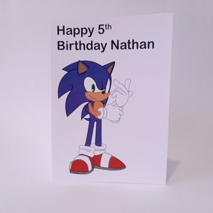 May include: A birthday card featuring Sonic the Hedgehog. The card has the text "Happy 5th Birthday Nathan" at the top. Sonic is depicted in his classic pose, with blue fur, red shoes, and a tan belly.
