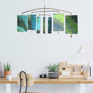 May include: A mobile made of eight rectangular pieces of painted glass, each with a different abstract design in shades of blue and green. The mobile is suspended from a metal rod and hangs from the ceiling.