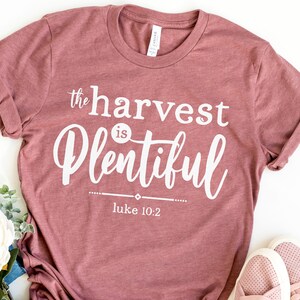 K&ouml;nnte beinhalten: Ein mauvefarbenes T-Shirt mit dem Text "the harvest is plentiful" und "luke 10:2" in wei&szlig;er Schrift.