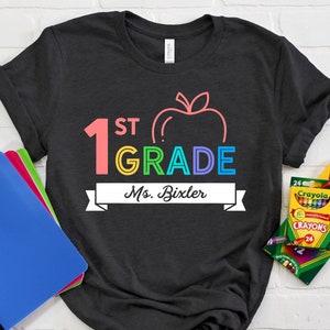 May include: A black t-shirt with a rainbow colored graphic of a large number 1, the word "ST" and the word "GRADE" with a red apple outline. The text "Ms. Bixler" is on a white banner below the graphic.