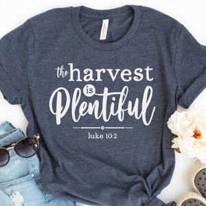 K&ouml;nnte beinhalten: Ein marineblaues T-Shirt mit wei&szlig;em Text, der "the harvest is plentiful" und "Luke 10:2" lautet.
