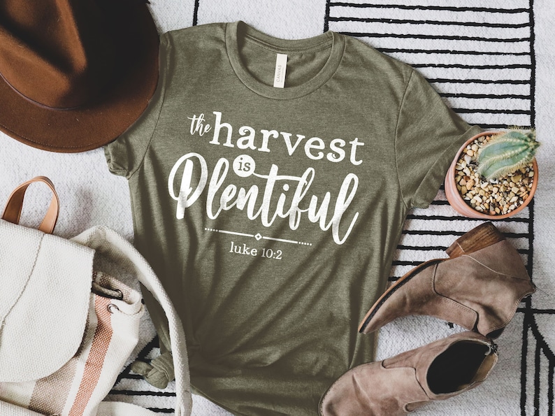 K&ouml;nnte beinhalten: Ein gr&uuml;nes T-Shirt mit wei&szlig;em Text, der "the harvest is plentiful" und "Luke 10:2" lautet.
