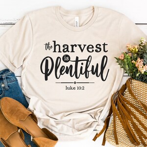 K&ouml;nnte beinhalten: Ein beiges T-Shirt mit dem Text "the harvest is plentiful" und "luke 10:2" in schwarzer Schrift.