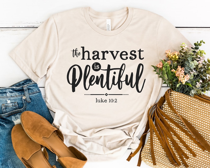 K&ouml;nnte beinhalten: Ein beiges T-Shirt mit dem Text "the harvest is plentiful" und "luke 10:2" in schwarzer Schrift.