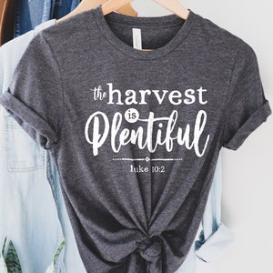 K&ouml;nnte beinhalten: Ein dunkelgraues T-Shirt mit wei&szlig;em Text, der "the harvest is plentiful" und "Luke 10:2" lautet.