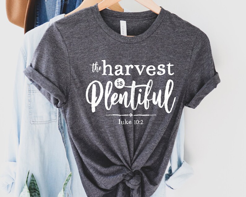 K&ouml;nnte beinhalten: Ein dunkelgraues T-Shirt mit wei&szlig;em Text, der "the harvest is plentiful" und "Luke 10:2" lautet.