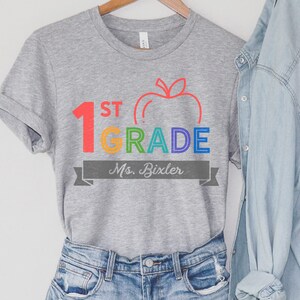May include: Gray t-shirt with a rainbow colored graphic that says "1st Grade" with an apple and the text "Ms. Bixler" in a gray banner.